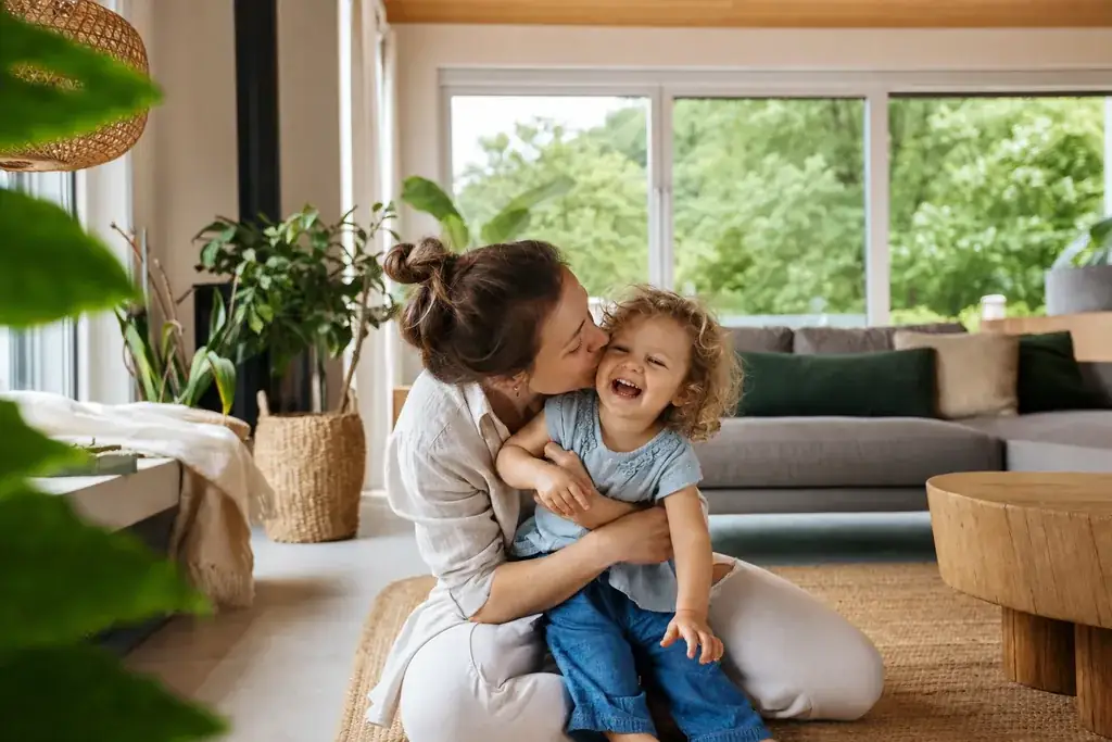 Symbolbild: eine Mutter herzt lachend ihr Kind in einem hellen, pflanzenreichen Wohnzimmer mit Blick ins Grüne, Sinnbild für sauberen Ökostrom und ein nachhaltiges Zuhause.